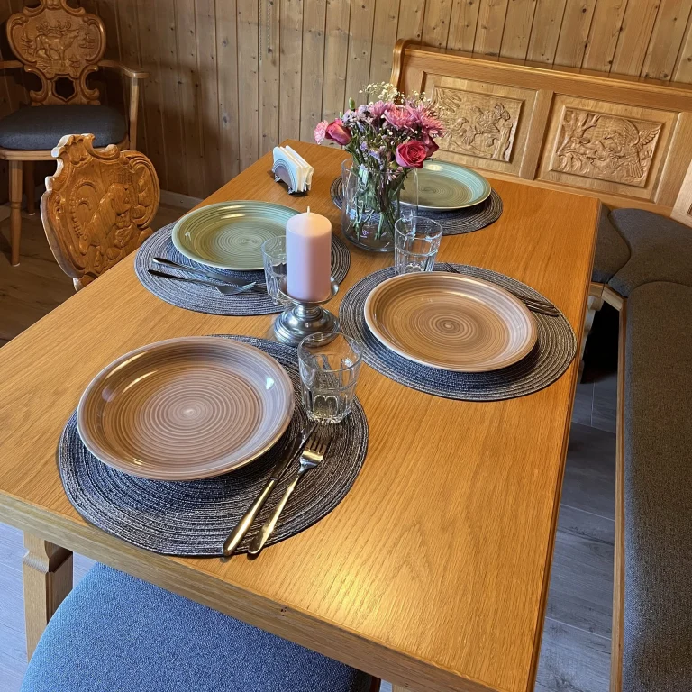 gedeckter-tisch-im-urigen-Esszimmer-mit-ansprechenen-geschirr Tisch mit gedecktem Geschirr, Blumenstrauß und Kerze in einem rustikalen Raum.