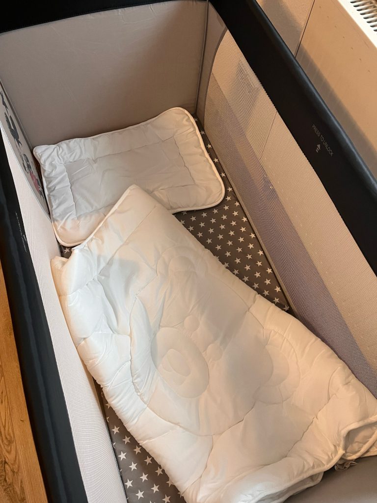 Casa Heckl Ferienwohnung Tutzing Traubing Starnberger See Weißes Babybett mit einer Decke und einem kleinen Kissen auf einem gepunkteten Untergrund.