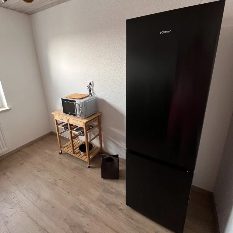 Kuechengeräte-Mikrowell-kuehl-gefrier-kombination-in-heller-moderner-kueche Schwarzer Kühlschrank neben einer Mikrowelle auf einem Holzregal in einem hellen Raum.