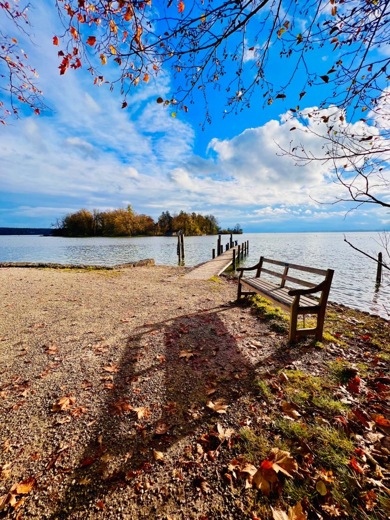 Tutzing Traubing Ferienwohnung Casa Heckl Blick auf einen ruhigen See mit einer Bank und buntem Herbstlaub im Vordergrund.