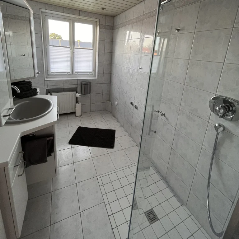 Helles Badezimmer mit Dusche, Waschbecken und großem Fenster. Bodenfliesen und Teppich. Helles Badezimmer mit Dusche, Waschbecken und großem Fenster. Bodenfliesen und Teppich.