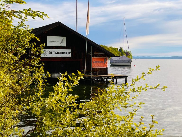 Tutzing Traubing Ferienwohnung Casa Heckl Schwarzes Bootshaus am Wasser, umgeben von grünen Pflanzen und Boote im Hintergrund.