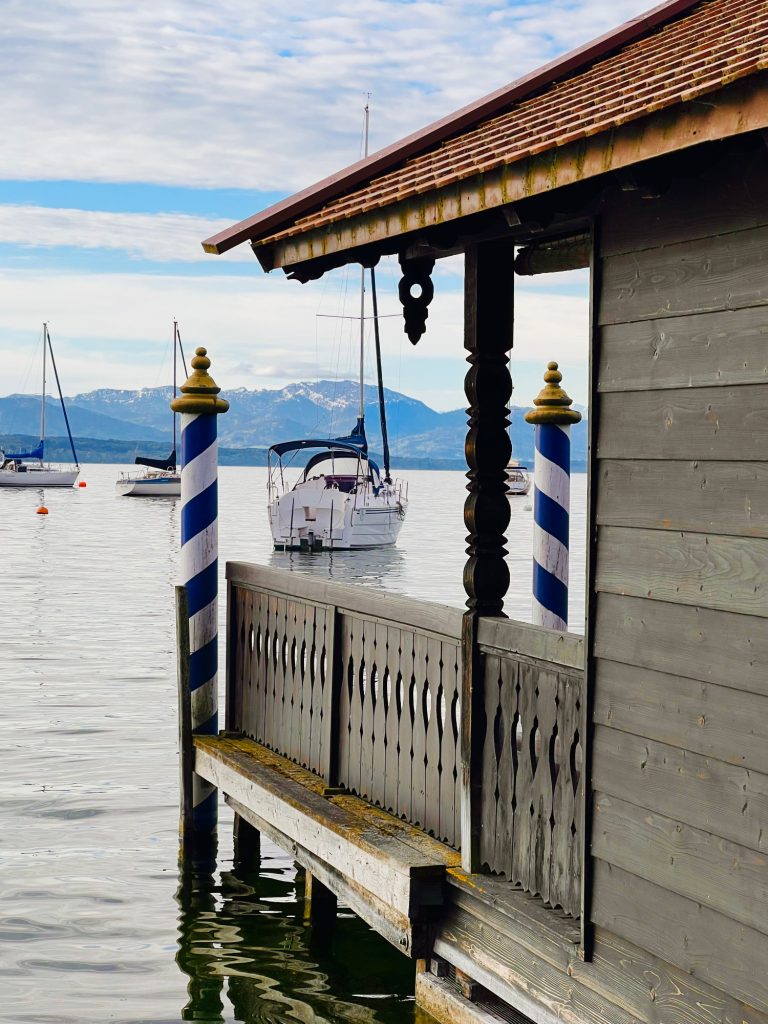 Tutzing Traubing Ferienwohnung Casa Heckl Holzsteg mit blauen Säulen, Boot im Wasser und Berge im Hintergrund.