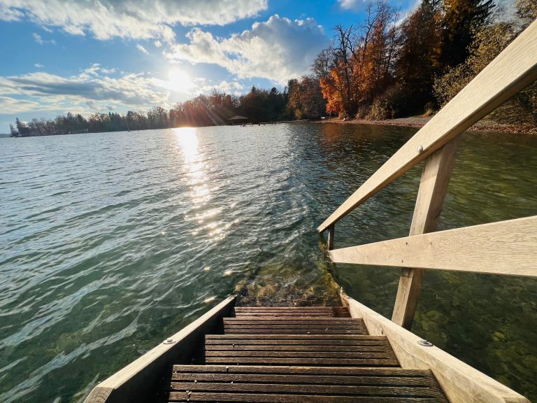 Tutzing Traubing Ferienwohnung Casa Heckl Holzstufen führen zu einem ruhigen Wasser mit Sonnenschein und herbstlichen Bäumen.
