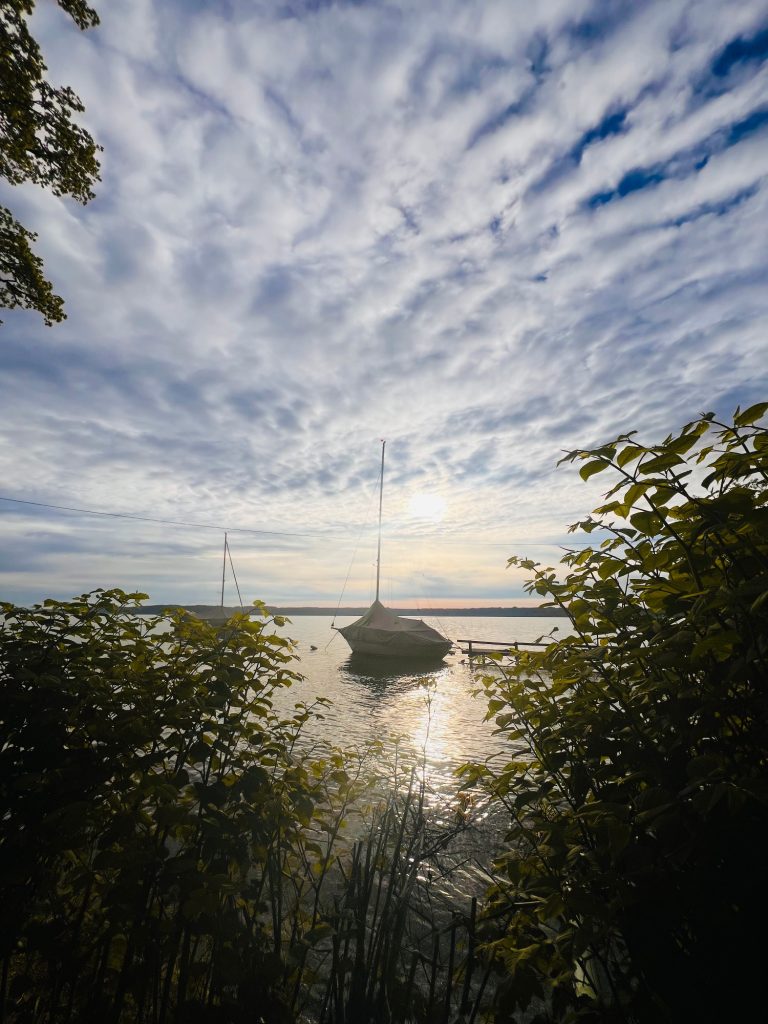 Tutzing Traubing Ferienwohnung Casa Heckl Sonnenuntergang über einem ruhigen Gewässer mit Segelboot und bewachsenen Ufern.