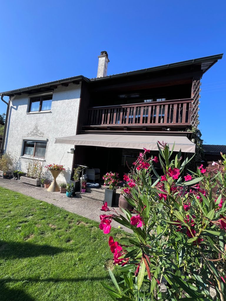 Casa Heckl Ferienwohnung Zweistöckiges Haus mit Balkon, umgeben von bunten Blumen und klarem blauen Himmel.