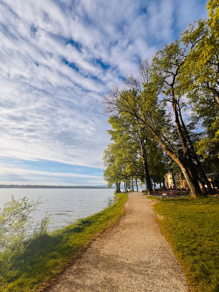 Tutzing Ferienwohnung Casa Heckl Uferweg mit Bäumen und Wasserblick unter einem bewölkten Himmel.