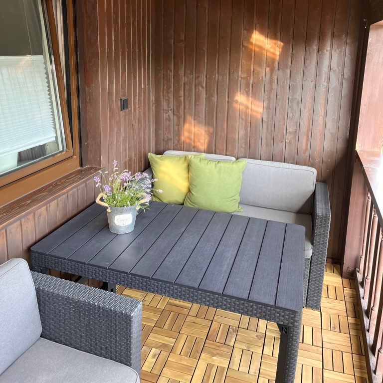 Casa Heckl Ferienwohnung Gemütlicher Balkon mit grauem Sofa, Kissen und Tisch, umgeben von Holzwänden.
