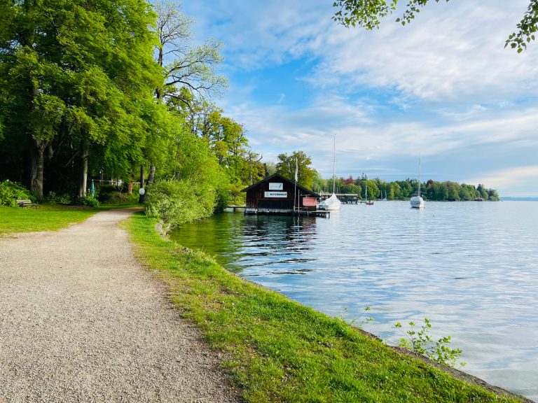 Tutzing Casa Heckl Ferienwohnung Uferweg entlang eines Sees mit einem Bootshaus und grünen Bäumen im Hintergrund.