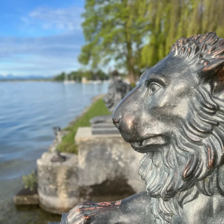 Löwenfigur am Wasser in Tutzing mit grünem Ufer und Blick auf den Starnberger See