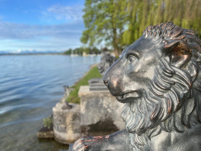 Tutzing Traubing Ferienwohnung Casa Heckl Bronzener Löwenkopf mit Blick auf einen ruhigen See und grüne Bäume im Hintergrund.