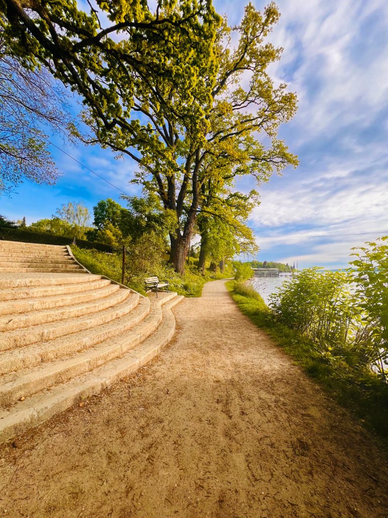 Tutzing Traubing Ferienwohnung Casa Heckl Uferweg mit Treppen und Bäumen am Wasser, unter blauem Himmel mit einigen Wolken.